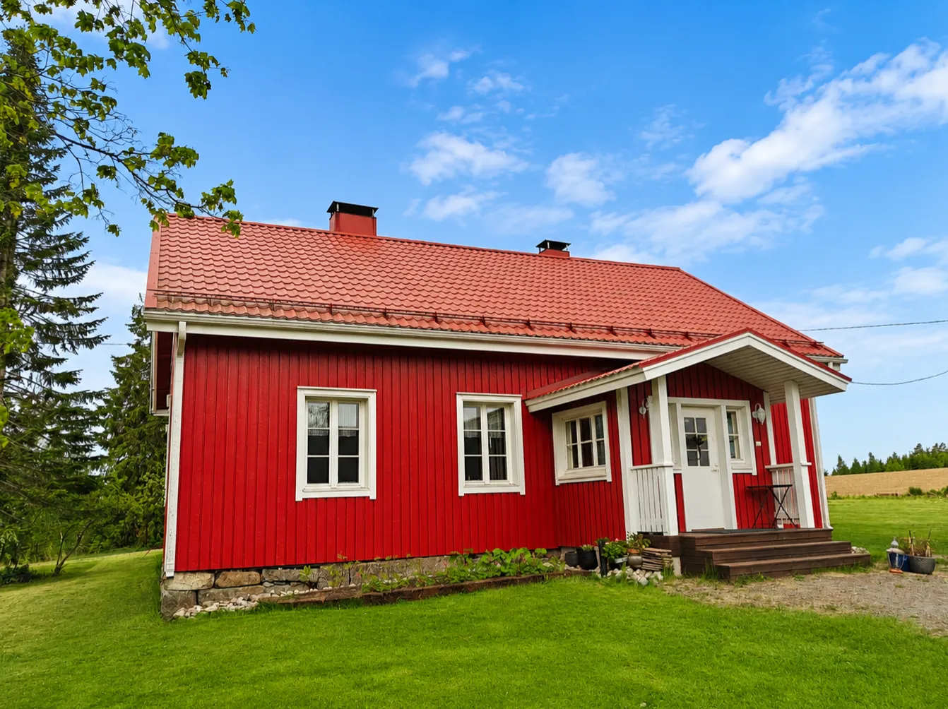 Punainen puutalo, jossa on punainen tiilikatto ja valkoinen julkisivuverhous, seisoo vihreällä nurmikolla sinisen taivaan alla.
