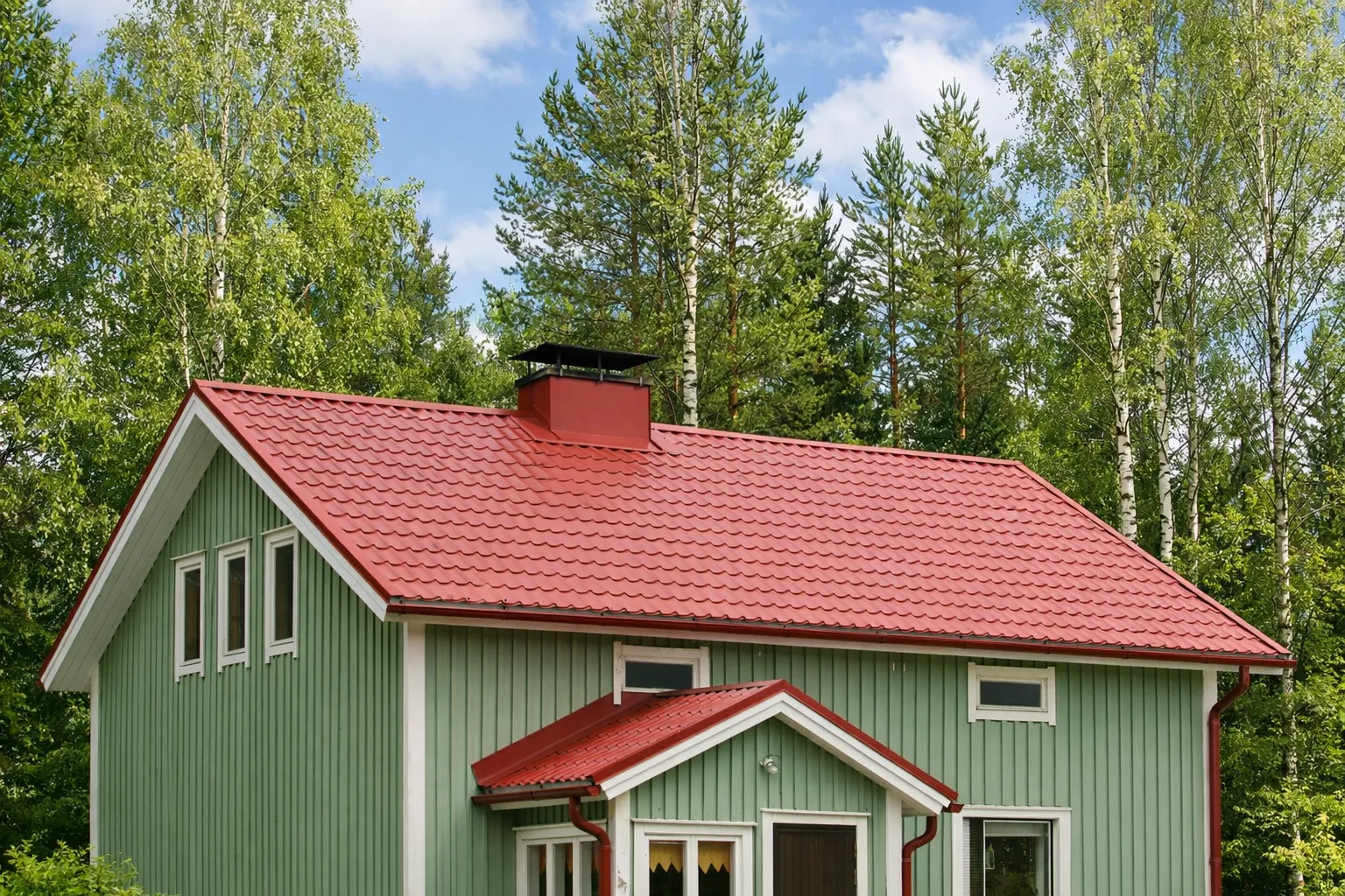 Vaaleanvihreä puutalo, jossa on punainen peltikatto ja julkisivu, seisoo puiden ympäröimänä sinisen taivaan alla.